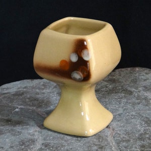 May include: A square-shaped ceramic planter with a brown and white glaze. The planter has a pedestal base and is decorated with brown and orange spots.