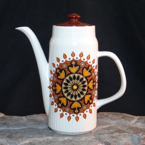 May include: A white ceramic coffee pot with a brown lid and a brown, orange and black geometric design. The pot has a rounded body and a spout on the side.