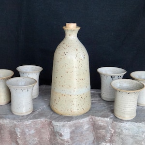 May include: A handmade ceramic decanter with a cork stopper, surrounded by six small drinking cups. The set is a speckled beige colour with a light brown band near the base. The cups have a flared rim.