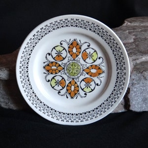 May include: A white ceramic plate with a black and white border and a colourful floral design in the centre. The design features orange, yellow, and green colours.