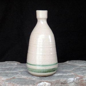 Peut inclure: Une bouteille en céramique blanche avec une bande verte autour du milieu. La bouteille a un fond arrondi et un col étroit.