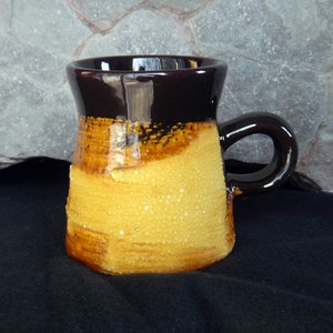 May include: A handcrafted ceramic mug. It features a dark brown rim and handle, blending into a textured yellow and brown body. The mug has a rounded handle and a glossy finish. Ideal for hot beverages.
