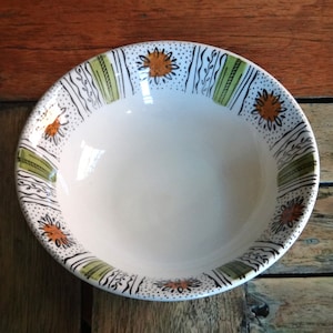 May include: A white ceramic bowl with a decorative rim featuring a repeating pattern of orange flowers, green stripes, and black and white abstract designs. The bowl is set against a rustic wooden surface.