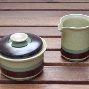 May include: A brown and cream ceramic sugar bowl with a lid and a matching creamer. The bowl and creamer have a brown, green and cream striped design.