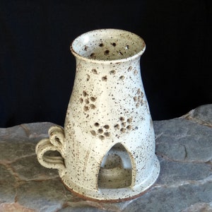 Peut inclure: Un bougeoir en céramique blanche avec un glaçage moucheté et un motif découpé. Le bougeoir a une poignée sur le côté et une petite ouverture au fond pour une bougie chauffe-plat.