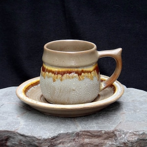 May include: A brown and beige ceramic coffee cup with a brown and yellow glaze sits on a matching saucer. The cup has a rounded handle.