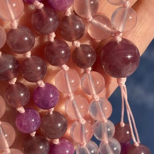 May include: A strand of pink and purple gemstone beads. The beads are round and have a smooth, polished finish. The beads are strung on a thin, white cord.
