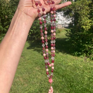 May include: A pink and white gemstone mala necklace with a tassel. The necklace is made of round beads and has a clear crystal bead at the end of the tassel.