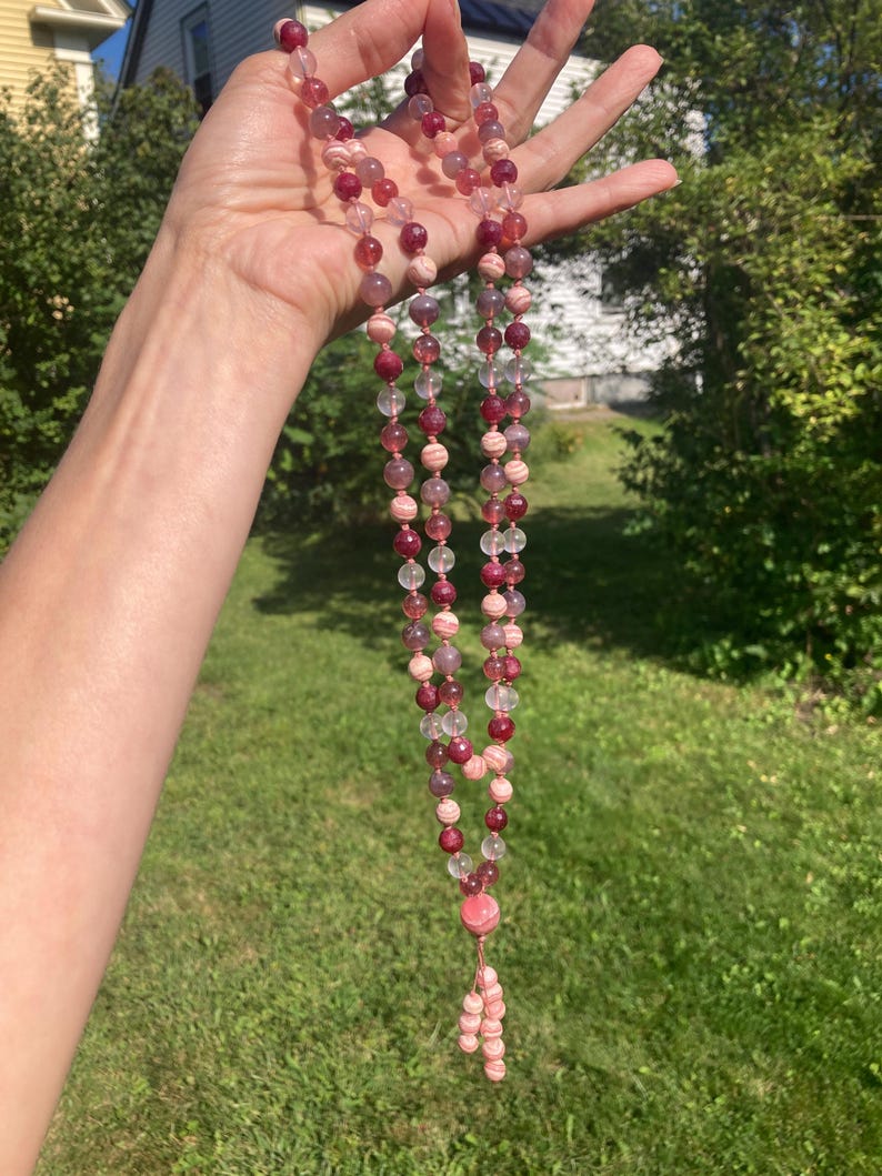 May include: A pink and white gemstone mala necklace with a tassel. The necklace is made of round beads and has a clear crystal bead at the end of the tassel.