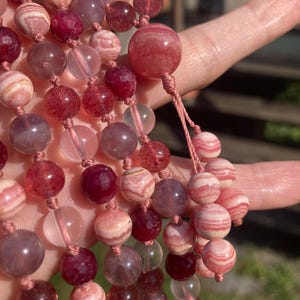 May include: A multi-strand necklace made with pink, red, and clear gemstone beads. The beads are round and have a smooth, polished finish. The necklace is strung on a thin, white cord.