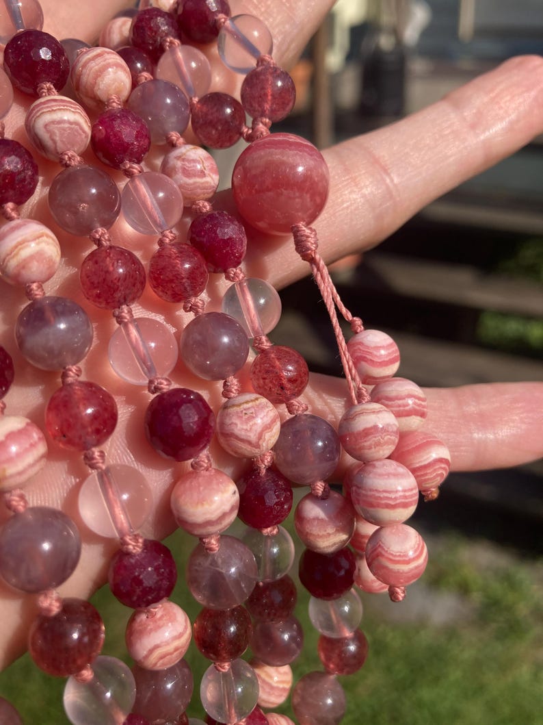 May include: A multi-strand necklace made with pink, red, and clear gemstone beads. The beads are round and have a smooth, polished finish. The necklace is strung on a thin, white cord.