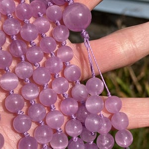 May include: A strand of light purple gemstone beads, likely amethyst, held in a hand. The beads are round and have a smooth, polished surface. The string is a matching light purple color. The background is blurred, suggesting an outdoor setting.
