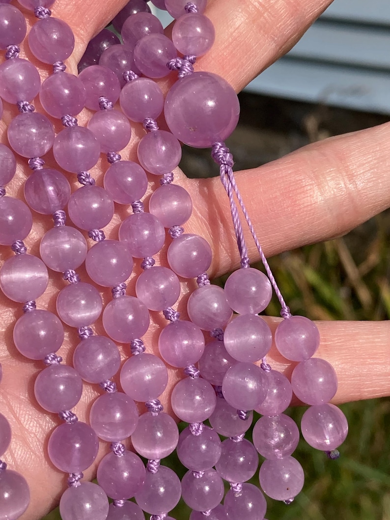 May include: A strand of light purple gemstone beads, likely amethyst, held in a hand. The beads are round and have a smooth, polished surface. The string is a matching light purple color. The background is blurred, suggesting an outdoor setting.