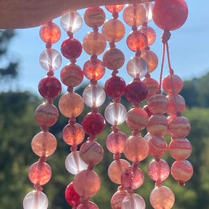 May include: A multi-strand necklace made of pink and white gemstone beads. The beads are round and have a smooth, polished finish. The necklace is strung on a thin, red cord.