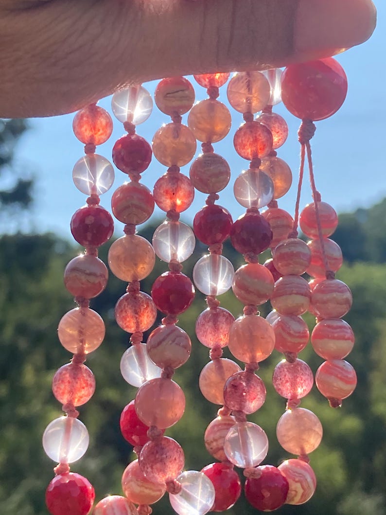 May include: A multi-strand necklace made of pink and white gemstone beads. The beads are round and have a smooth, polished finish. The necklace is strung on a thin, red cord.