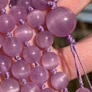 May include: Close-up of a strand of amethyst beads. The beads are a soft purple color and are round, with a smooth, polished surface. The beads are strung together with a matching purple cord, knotted between each bead. The background is blurred.
