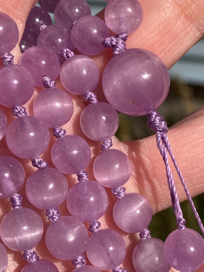 May include: Close-up of a strand of amethyst beads. The beads are a soft purple color and are round, with a smooth, polished surface. The beads are strung together with a matching purple cord, knotted between each bead. The background is blurred.