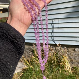 May include: A long, beaded necklace with light purple, round beads. The necklace is held up against a blue and white background. The necklace has a tassel of smaller beads at the end.