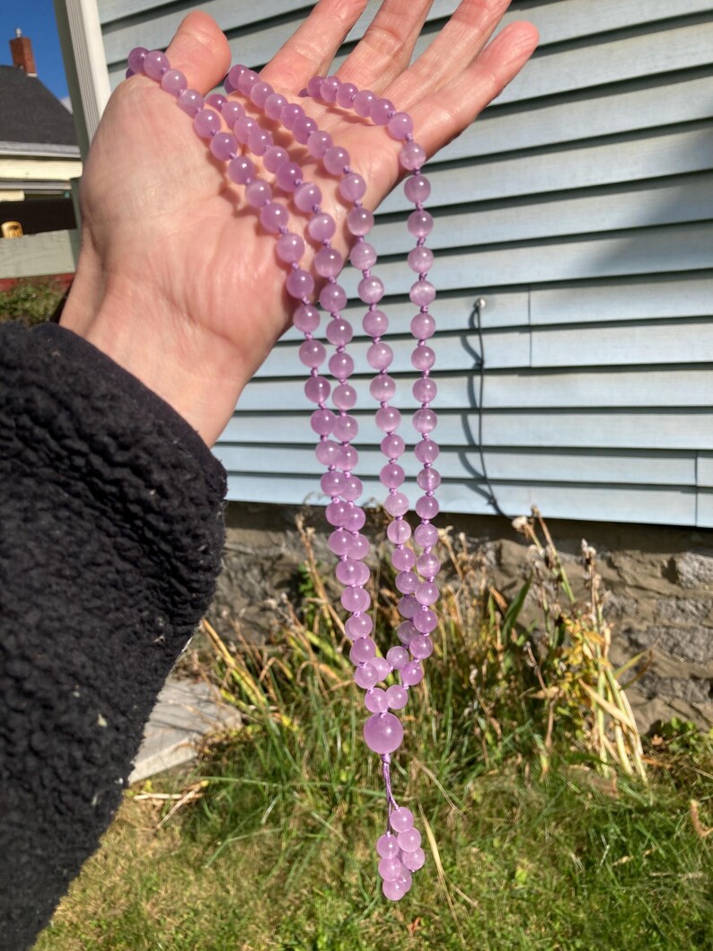 May include: A long, beaded necklace with light purple, round beads. The necklace is held up against a blue and white background. The necklace has a tassel of smaller beads at the end.