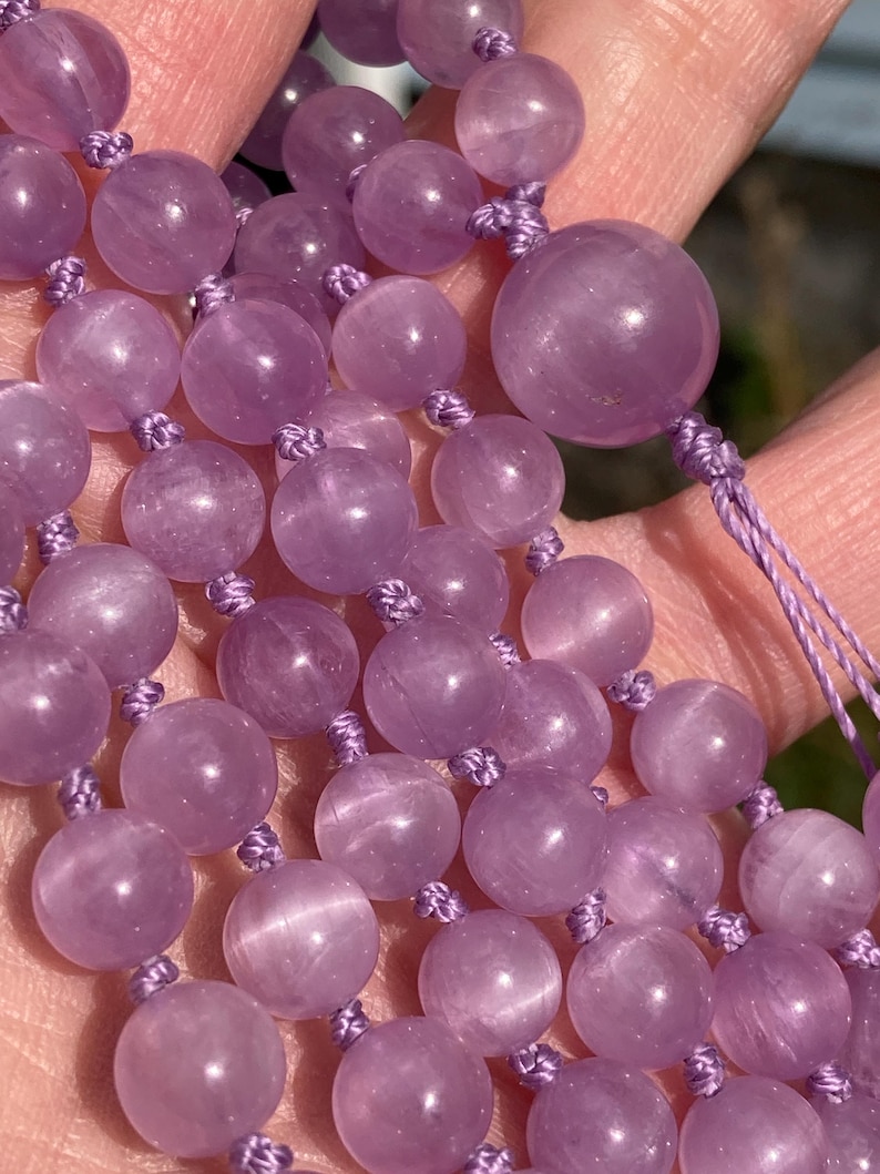 May include: Close-up of a lavender-colored beaded necklace. The beads are round and strung together with matching lavender-colored cord. The beads have a smooth, polished surface and vary slightly in size.