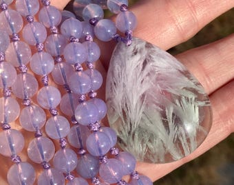 May include: A purple and white crystal necklace with a large, teardrop-shaped pendant. The pendant is made of clear crystal with white, feathery inclusions.