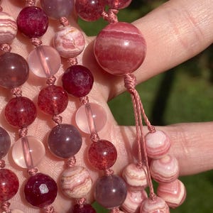 May include: A strand of pink and red gemstone beads with a knotted tassel. The beads are round and have a smooth, polished finish. The beads are strung on a thin, brown cord.