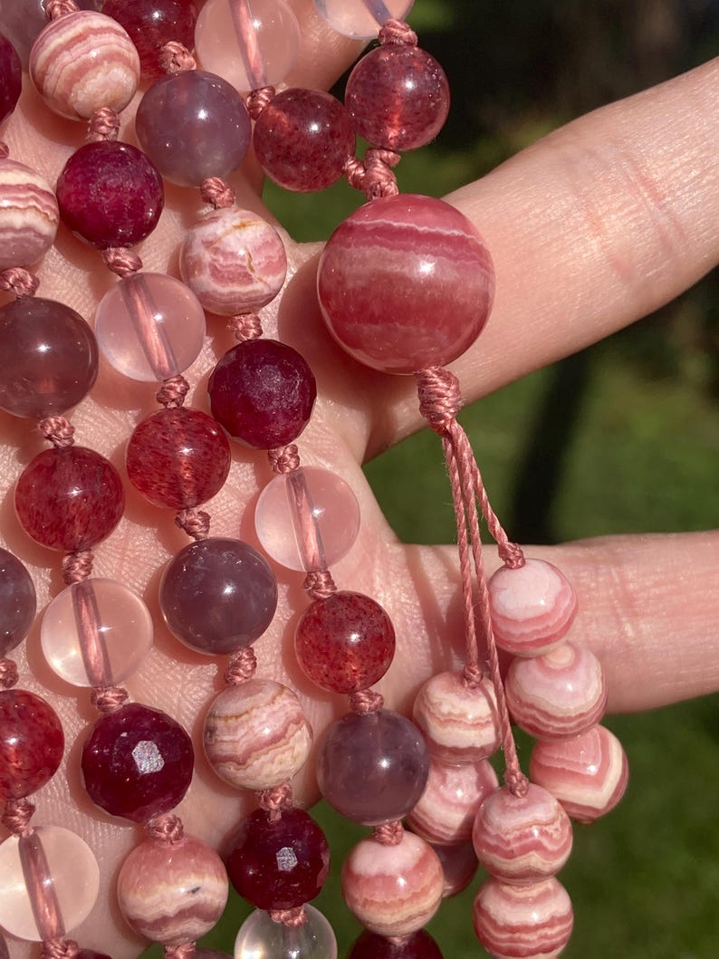 May include: A strand of pink and red gemstone beads with a knotted tassel. The beads are round and have a smooth, polished finish. The beads are strung on a thin, brown cord.