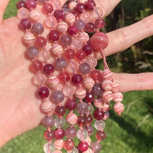 May include: A multi-strand necklace made with pink, white, and clear beads. The beads are made of various materials, including agate, quartz, and jasper. The necklace is strung on a pink cord.