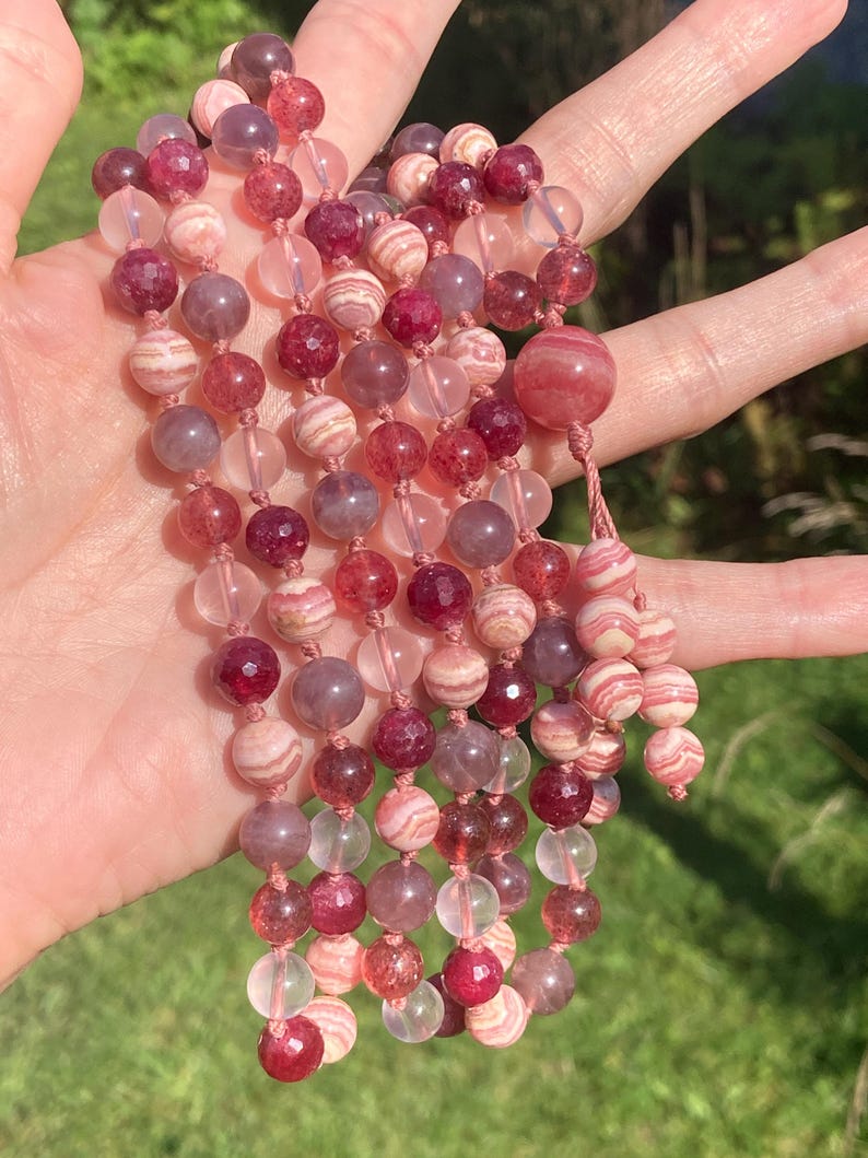May include: A multi-strand necklace made with pink, white, and clear beads. The beads are made of various materials, including agate, quartz, and jasper. The necklace is strung on a pink cord.