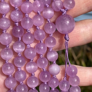 May include: Close-up of a strand of amethyst beads. The beads are a soft lavender color and are strung on a matching purple cord. The beads are round and smooth, with a slightly translucent appearance. The mala beads are a spiritual accessory.