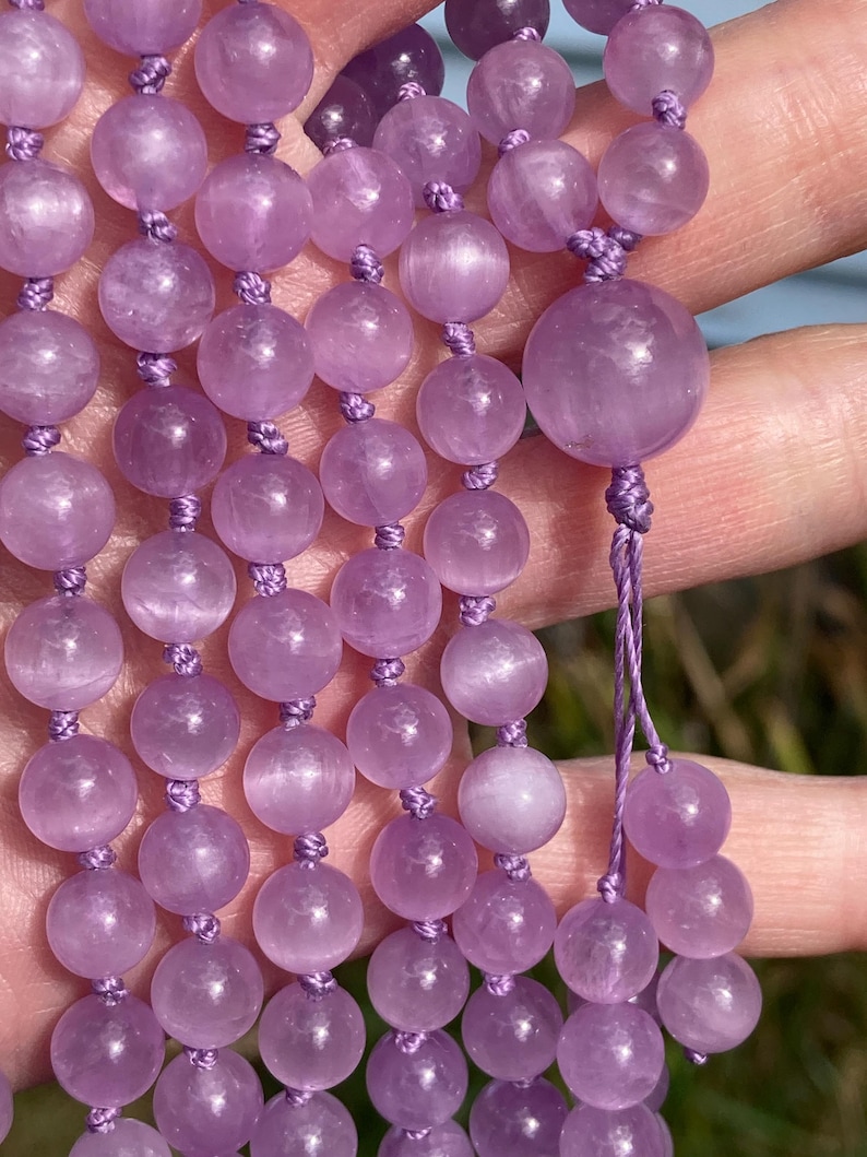 May include: Close-up of a strand of amethyst beads. The beads are a soft lavender color and are strung on a matching purple cord. The beads are round and smooth, with a slightly translucent appearance. The mala beads are a spiritual accessory.