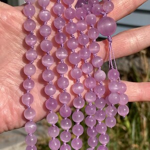 May include: A strand of light purple gemstone beads, likely amethyst, strung on a matching cord. The beads are round and smooth, with a subtle sheen. The necklace is held up, showcasing the color and texture of the beads.