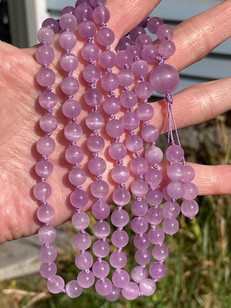 May include: A strand of light purple gemstone beads, likely amethyst, strung on a matching cord. The beads are round and smooth, with a subtle sheen. The necklace is held up, showcasing the color and texture of the beads.