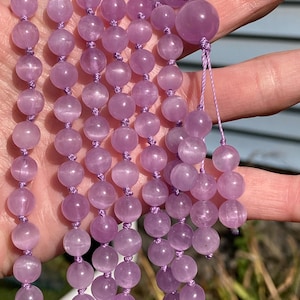May include: A strand of light purple gemstone beads, likely amethyst, strung together with matching purple thread. The beads are round and smooth, with a slightly translucent appearance. The mala beads are held in a hand.