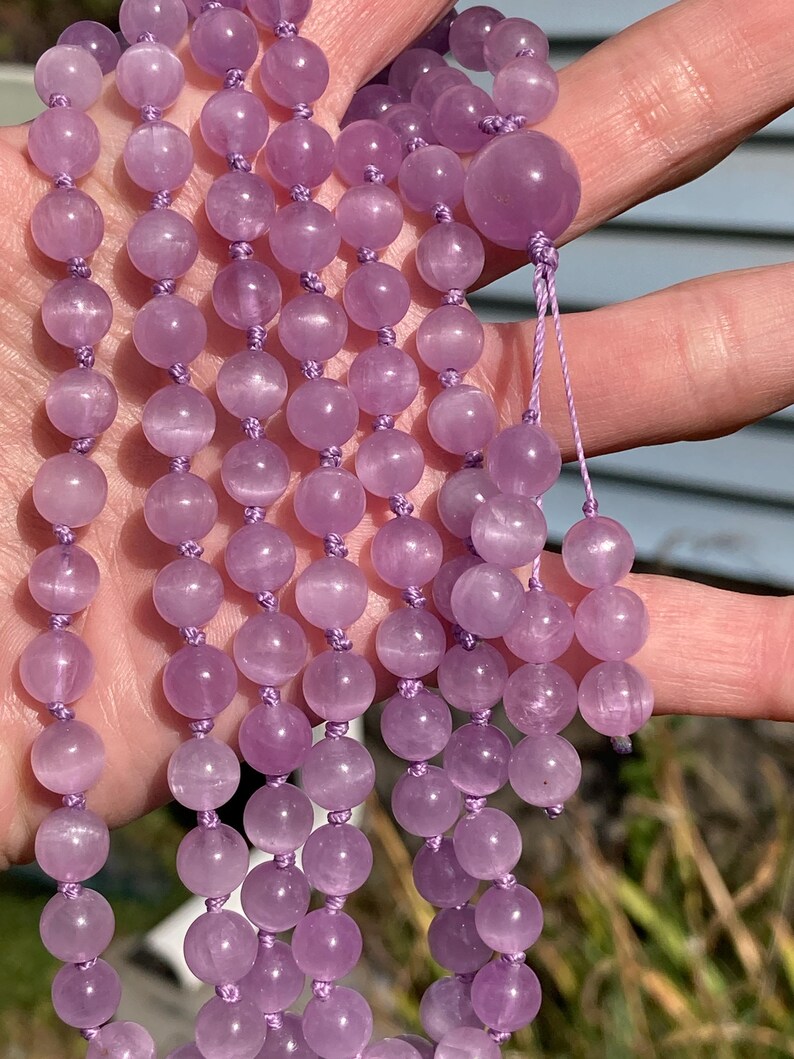 May include: A strand of light purple gemstone beads, likely amethyst, strung together with matching purple thread. The beads are round and smooth, with a slightly translucent appearance. The mala beads are held in a hand.