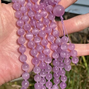 May include: A strand of light purple gemstone beads, likely amethyst, strung together with matching thread. The beads are round and smooth, with a soft, translucent appearance. The necklace is held in a hand, showcasing its length and color.