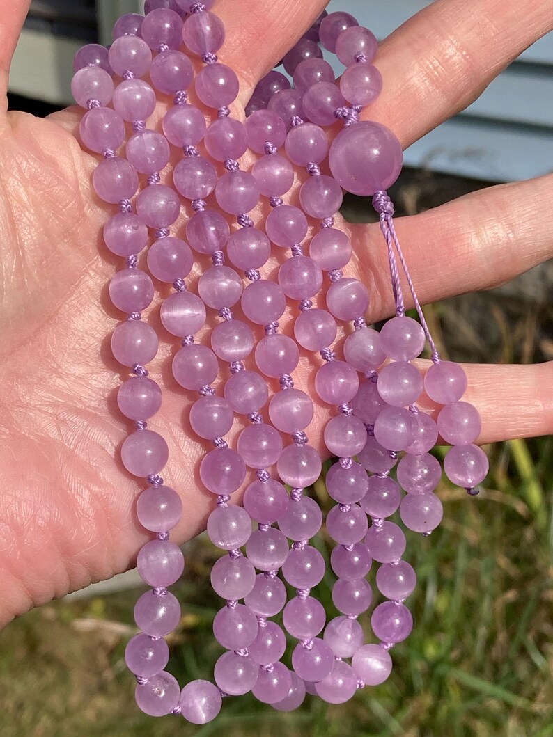 May include: A strand of light purple gemstone beads, likely amethyst, strung together with matching thread. The beads are round and smooth, with a soft, translucent appearance. The necklace is held in a hand, showcasing its length and color.