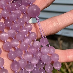 May include: A strand of lavender-colored gemstone beads, likely amethyst, strung on a matching cord. The beads are round and smooth, with a polished finish. The necklace is held in a hand, showcasing the color and texture of the beads.