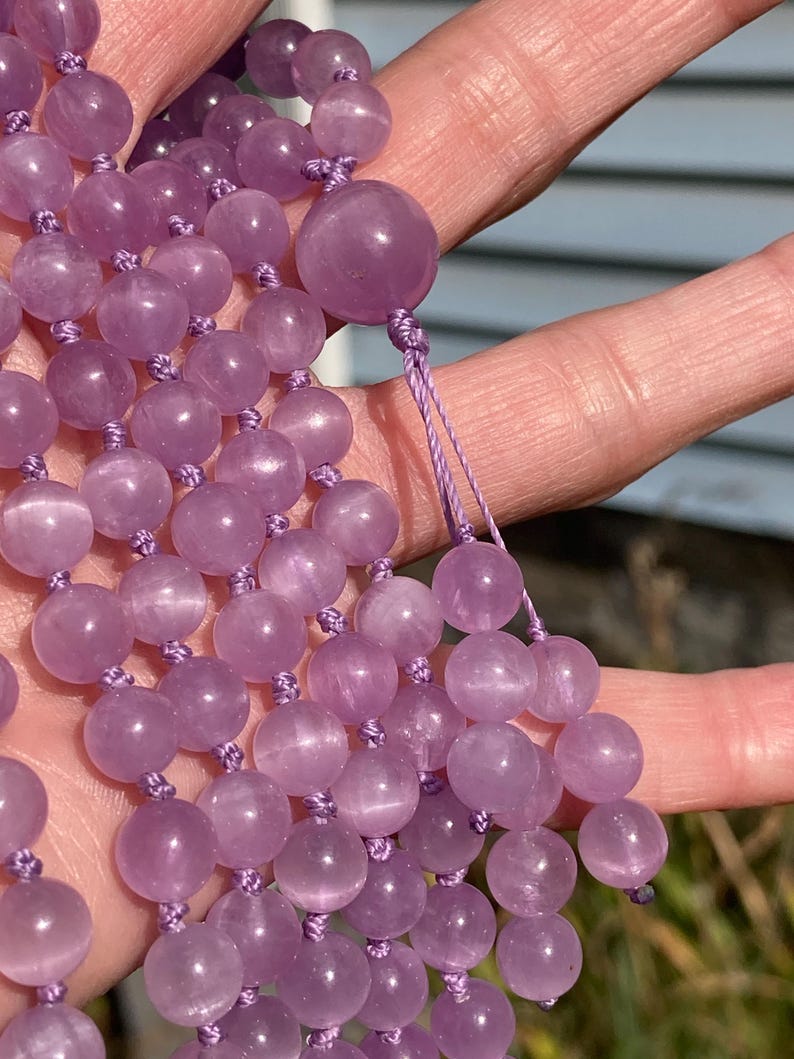 May include: A strand of lavender-colored gemstone beads, likely amethyst, strung on a matching cord. The beads are round and smooth, with a polished finish. The necklace is held in a hand, showcasing the color and texture of the beads.