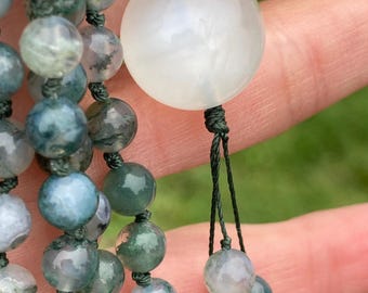 May include: A beaded necklace featuring numerous round moss agate beads in shades of green, gray, and white, strung on dark green cord. A single, large, smooth, translucent white stone bead serves as a focal point, accented by a dangling bead.