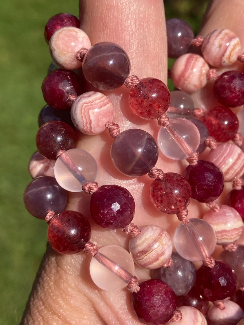 May include: A close-up of a multi-strand beaded necklace with pink, red, and clear beads. The beads are made of various materials, including gemstone and glass.