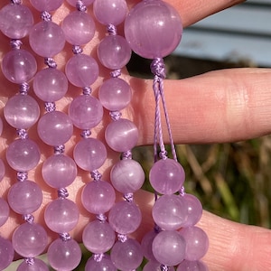 May include: Close-up of a strand of lavender-colored gemstone beads. The beads are round and strung together with matching lavender-colored cord. The beads have a smooth, polished surface and are knotted between each bead. The background is blurred.