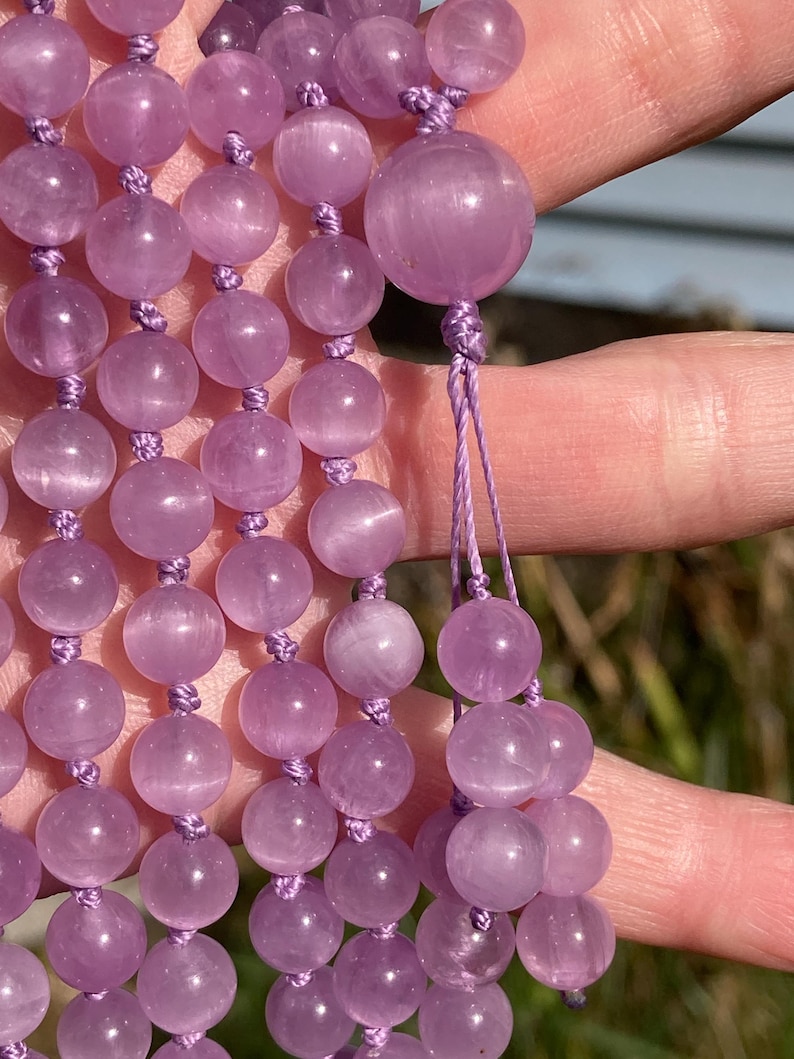 May include: Close-up of a strand of lavender-colored gemstone beads. The beads are round and strung together with matching lavender-colored cord. The beads have a smooth, polished surface and are knotted between each bead. The background is blurred.