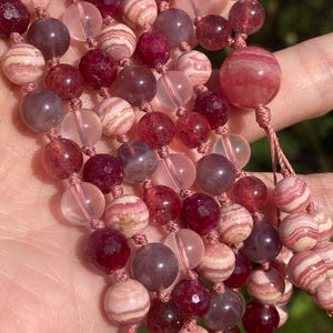 May include: A multi-strand necklace made of pink, red, and clear beads. The beads are round and have a smooth, polished finish. The necklace is strung on a thin, pink cord.
