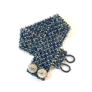 May include: A wide cuff bracelet crafted from a dense weave of small, dark blue and metallic silver seed beads. The bracelet features two round, off-white mother-of-pearl buttons as a closure mechanism, paired with two matching beaded loops.
