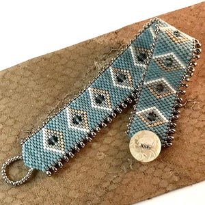 May include: A wide beaded bracelet featuring a geometric diamond pattern in shades of light blue, cream, and dark gray. The edges are lined with metallic silver beads, and the closure consists of a round, white, floral-patterned button.