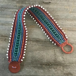 May include: A beaded bracelet featuring rows of teal, blue, and terracotta beads with a white beaded fringe along the edges. The bracelet includes a circular terracotta-colored button closure at one end and a matching beaded loop at the other end.