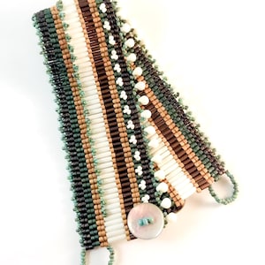 May include: A wide cuff bracelet featuring horizontal rows of tubular beads in white, brown, and tan, accented by vertical columns of dark green and teal seed beads. The edges are finished with teal picot loops and a single iridescent shell button.