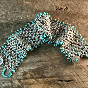 May include: A wide woven cuff bracelet featuring a grid pattern of metallic bronze and silver square beads, accented with small turquoise blue seed beads along the edges and two circular white buttons used as fasteners at the ends.