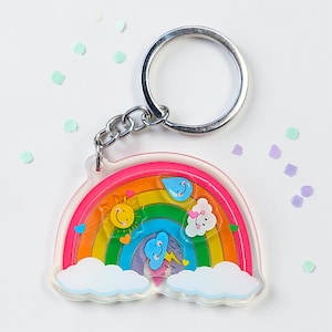 Puede incluir: Un llavero de acrílico transparente con un diseño de arcoíris colorido que incluye un sol sonriente, una gota de lluvia azul, una nube blanca y una nube de tormenta azul con un rayo amarillo. El dije está unido a una argolla de metal plateado.